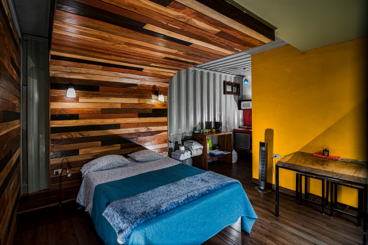 Interior del Studio Maracuyá con cama queen, techo de maderas, pared amarilla y zona de cocina